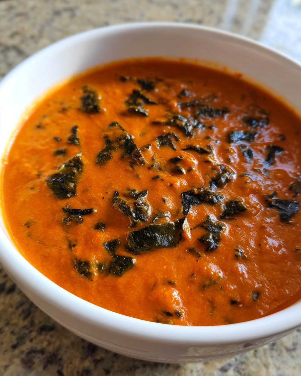 Close-up of creamy orange tomato basil soup topped with dark green wilted greens in a white bowl.