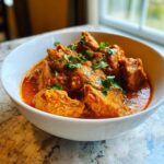 A close-up of rich, orange-red Crockpot Butter Chicken served in a white bowl and garnished with fresh cilantro.