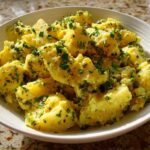 A white bowl filled with bright yellow CRUSHED POTATO SALAD, generously coated in a dressing with capers and fresh parsley.