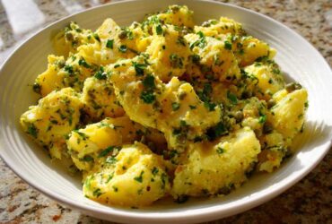 A white bowl filled with bright yellow CRUSHED POTATO SALAD, generously coated in a dressing with capers and fresh parsley.
