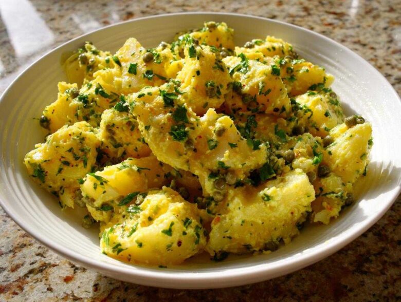 A white bowl filled with bright yellow CRUSHED POTATO SALAD, generously coated in a dressing with capers and fresh parsley.