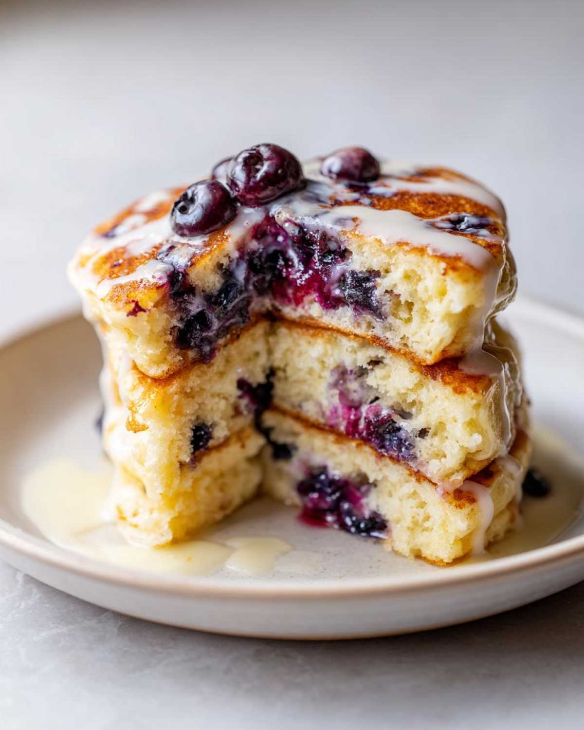 Close-up of a stack of fluffy Delicious Blueberry Pancakes cut in half, drizzled with glaze and topped with fresh blueberries.