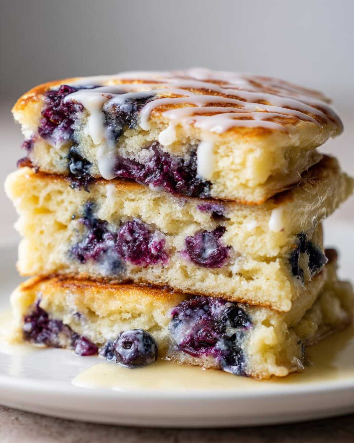 Close-up of a stack of three fluffy Delicious Blueberry Pancakes oozing with purple berries and drizzled with white glaze.