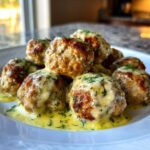 A pile of golden brown Easy Creamy Greek Chicken Meatballs covered in a rich, creamy yellow sauce and sprinkled with fresh dill.