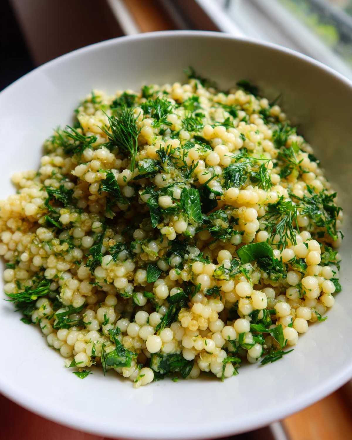 Close-up of a white bowl filled with Easy One-Pot Creamy Lemon Herb Couscous topped generously with fresh green herbs like dill and parsley.