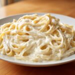 Close-up of fettuccine pasta generously coated in rich, white Creamy Alfredo Sauce, seasoned with black pepper.