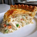Close-up of a thick slice of chicken pot pie recipe showing creamy filling, shredded chicken, peas, and carrots under a flaky golden crust.