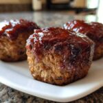 Close-up of three perfectly portioned mini meatloaf items topped with a thick, shiny barbecue glaze, resting on a white plate.
