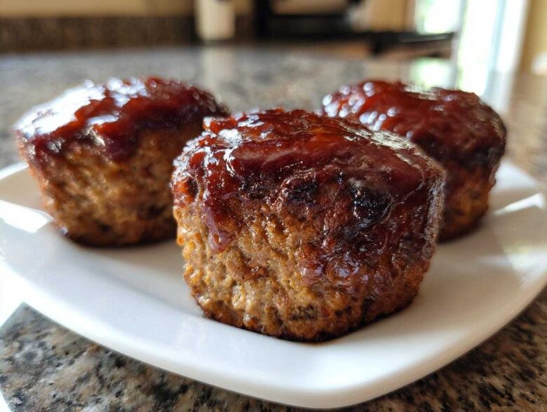 Close-up of three perfectly portioned mini meatloaf items topped with a thick, shiny barbecue glaze, resting on a white plate.