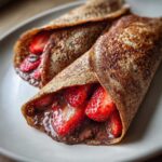 Close-up of two rolled Gluten-Free Chocolate Strawberry Crepes filled with chocolate spread and fresh sliced strawberries on a light plate.