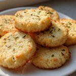 A stack of golden Gluten-Free Lemon Thyme Shortbread Cookies sprinkled with dried herbs on a white plate.