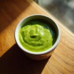 A small white bowl filled with thick, bright green Green Goddess Salad Dressing, sitting on a sunny wooden surface.