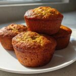 Four golden-brown High Protein Banana Muffins stacked slightly on a white plate, backlit by sunlight.