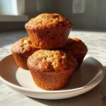A stack of four golden-brown High Protein Banana Muffins served on a light-colored plate.