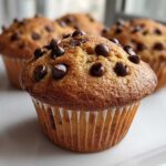 A close-up of a perfectly domed Homemade Chocolate Chip Muffins, studded with melted chocolate chips, sitting on a white plate.