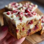 A hand holds up a thick, moist square of cranberry bliss bars topped with white chocolate frosting and festive sprinkles.