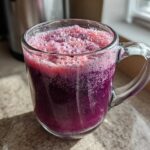 Close-up of a vibrant, deep purple Iced Blueberry Lemonade Slush with frothy pink foam in a clear glass mug.