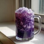 A refreshing glass mug filled high with vibrant purple Iced Blueberry Lemonade Slush, sitting on a sunny countertop.