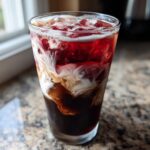 A tall glass showing distinct layers of dark cold brew, creamy white vanilla swirl, and bright red cherry syrup in an Iced Cherry Vanilla Cold Brew.