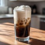 A tall glass filled with an Iced Coconut Mocha with Cold Foam, showing dark coffee mixing with foam.