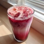 A tall glass filled with a vibrant, layered Iced Raspberry Vanilla Chai, topped with ice and foam.