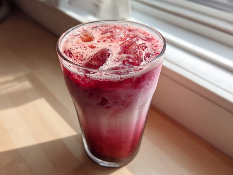 A tall glass filled with a vibrant, layered Iced Raspberry Vanilla Chai, topped with ice and foam.