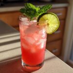 A tall glass filled with a vibrant pink Iced Watermelon Lime Cooler, ice, and garnished with fresh mint and a lime wheel.