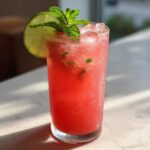 A tall glass filled with a vibrant red Iced Watermelon Lime Cooler, garnished with fresh mint and a lime wheel.