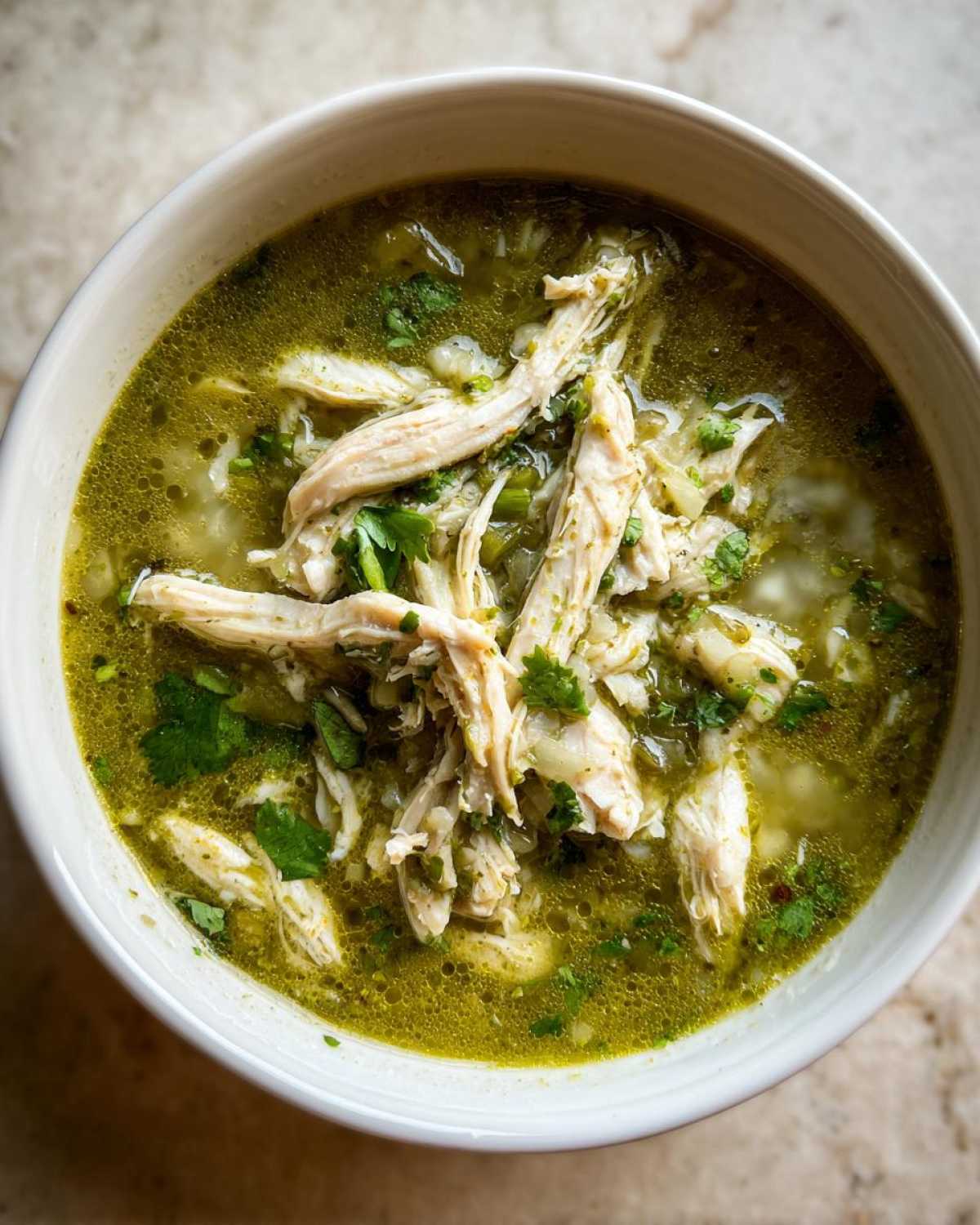Overhead shot of a white bowl filled with vibrant green Instant Pot Chicken Pozole Verde, topped generously with shredded chicken and cilantro.