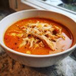 A close-up of a white bowl filled with rich, orange Instant Pot Creamy Chicken Enchilada Soup, topped with shredded chicken and cheese.