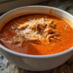 A close-up of a white bowl filled with rich, orange Instant Pot Creamy Chicken Enchilada Soup topped with shredded chicken and melted cheese.