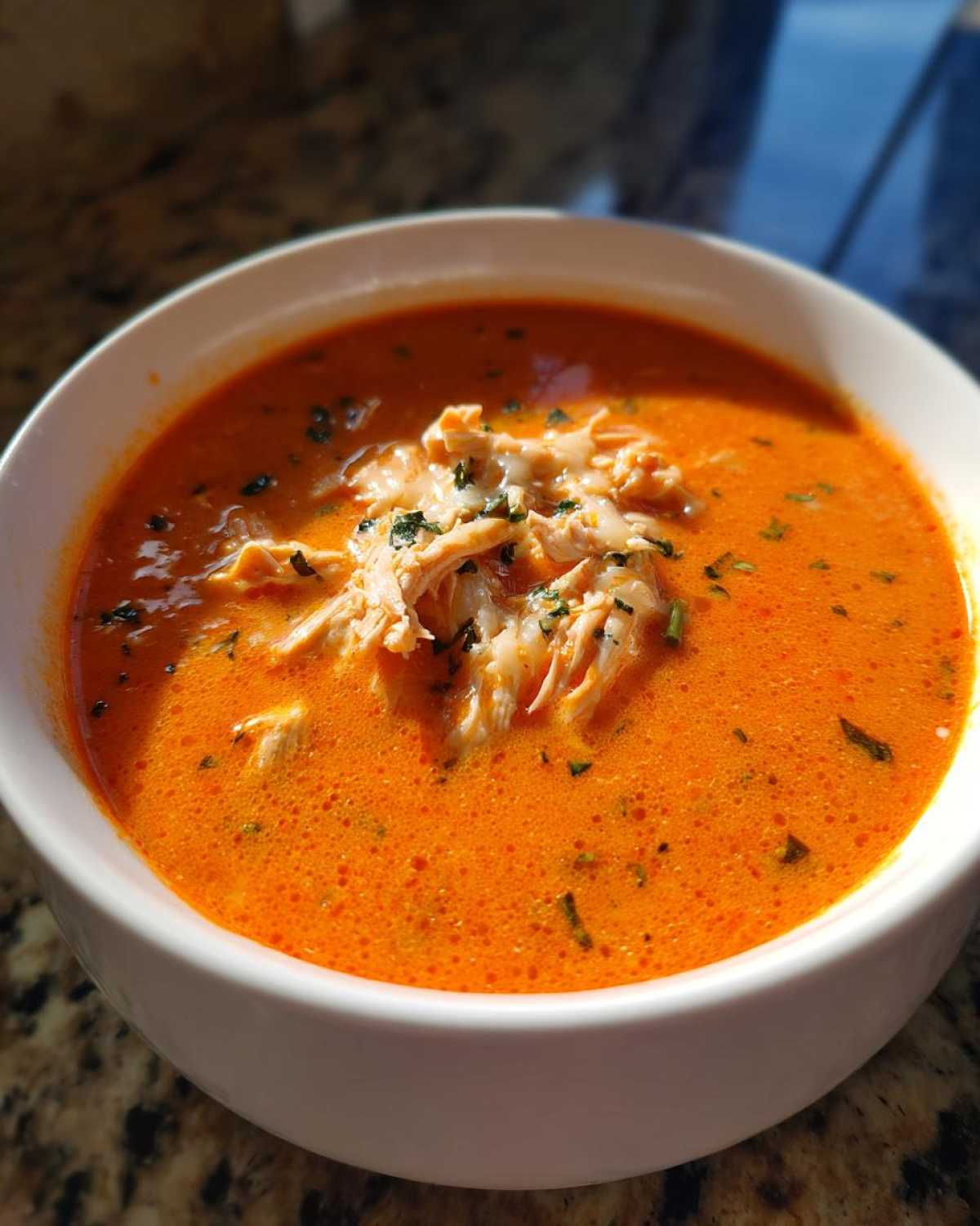Close-up of a white bowl filled with vibrant orange Instant Pot Creamy Chicken Enchilada Soup, topped with shredded chicken and herbs.