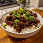 A close-up of a white bowl filled with white rice topped generously with saucy Instant Pot Korean Beef and garnished with sliced green onions and sesame seeds.