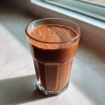 A glass filled with a thick, dark brown Keto Chocolate Strawberry Smoothie sitting on a white counter near a window.