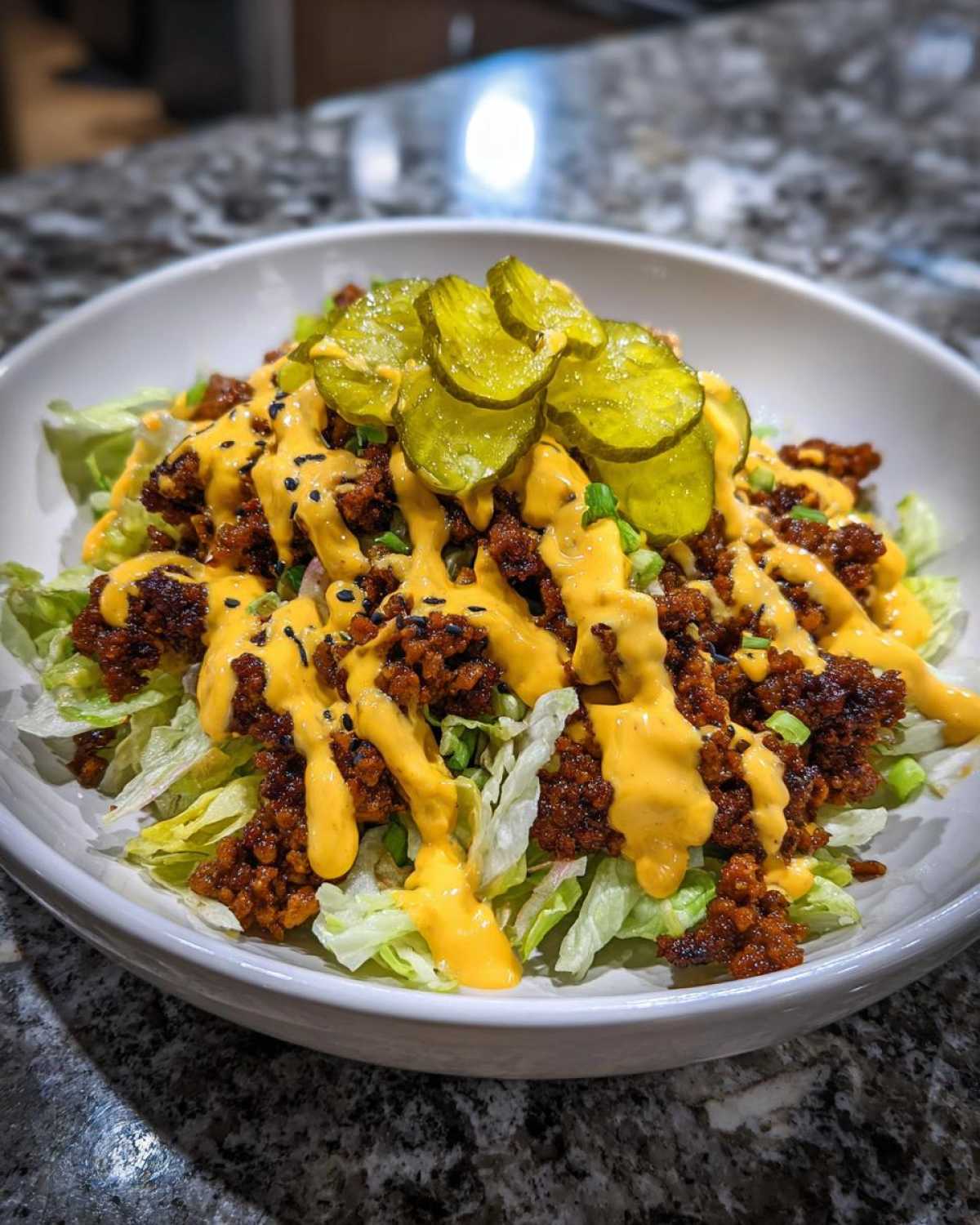 A close-up of a big mac salad featuring lettuce, seasoned ground meat, bright yellow sauce drizzle, and pickle slices on top.