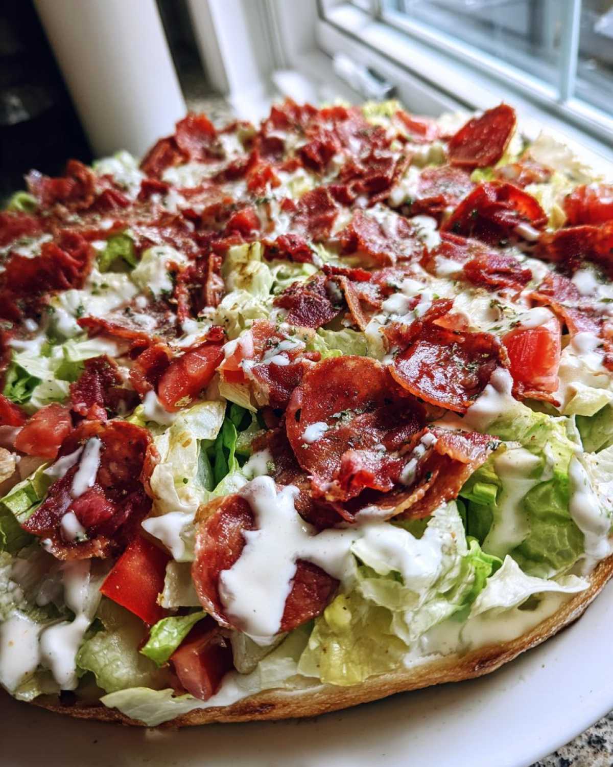 Close-up of a loaded grinder salad piled high on bread with lettuce, tomatoes, crispy meat, and creamy dressing.