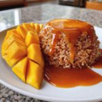 A serving of brown mango sticky rice topped with rich caramel sauce, next to perfectly sliced fresh mango.