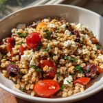 A close-up bowl of vibrant Mediterranean farro salad featuring whole farro grains, cherry tomatoes, Kalamata olives, and crumbled feta cheese.