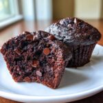 Close-up of two rich, dark chocolate muffins, one cut in half showing a moist crumb and chocolate chips, highlighting the texture of the chocolate muffins.
