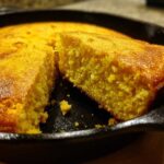 A close-up view of moist cornbread, with one slice cut and slightly pulled away in a black cast iron skillet.