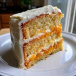 A close-up of a moist slice of Creamsicle Cake showing layers of yellow cake, bright orange filling, and white frosting.