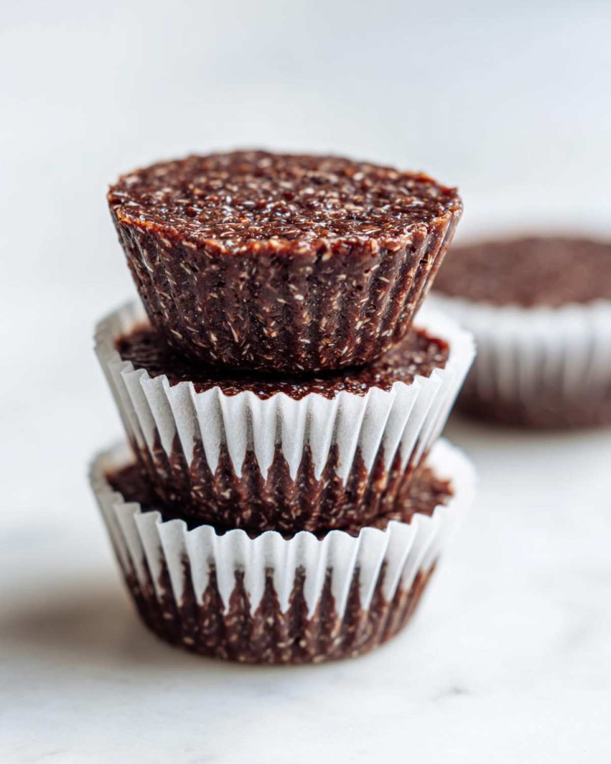 A stack of three dark, textured No-Bake Chocolate Coconut Protein Cups in white paper liners on a light surface.