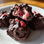 A close-up of several No-Bake Chocolate Strawberry Yogurt Clusters piled on a white plate, one is broken open showing the pink yogurt filling.