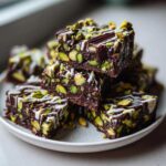 A stack of rich, dark No-Bake Pistachio Chocolate Fudge Squares topped with white chocolate drizzle and whole pistachios.