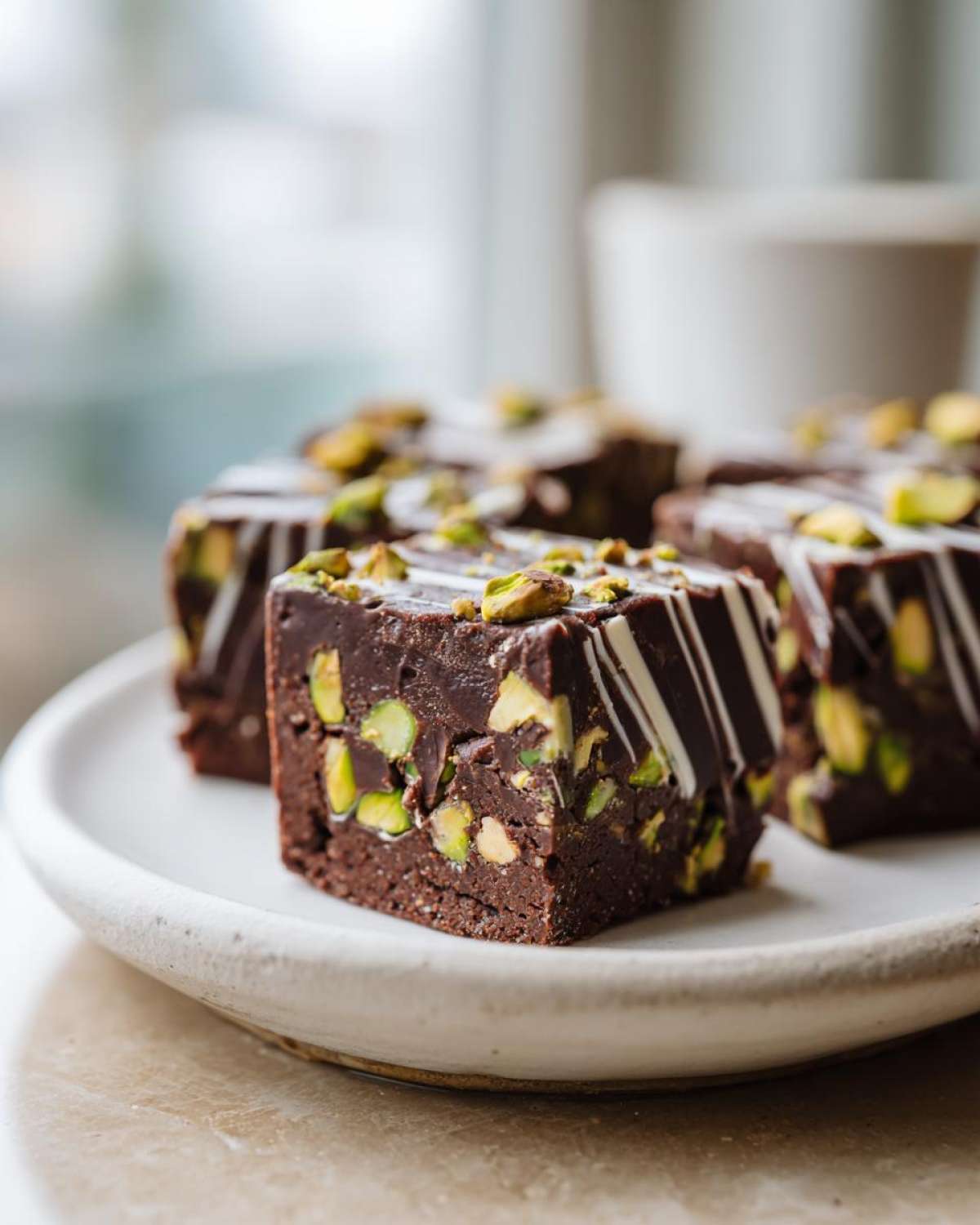 A close-up of rich, dark No-Bake Pistachio Chocolate Fudge Squares topped with white chocolate drizzle and chopped pistachios.