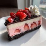 A decadent slice of No-Bake Strawberry Chocolate Cheesecake featuring layers of pink and white filling, whole strawberries, and a dark chocolate crust.