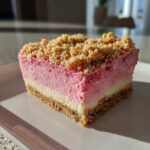 A close-up of a square slice of No Bake Strawberry Crunch Cheesecake showing graham cracker crust, white layer, pink strawberry layer, and crunchy topping.