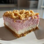A square slice of No Bake Strawberry Crunch Cheesecake showing graham cracker crust, white layer, pink layer, and crunchy topping.