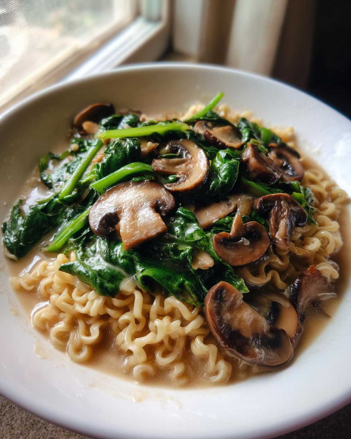 Close-up of One-Pot Creamy Mushroom Spinach Ramen featuring wavy noodles in a light sauce topped with sautéed mushrooms and wilted spinach.