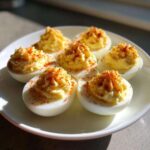 Six perfectly piped deviled eggs dusted with paprika, served on a white plate in bright sunlight.